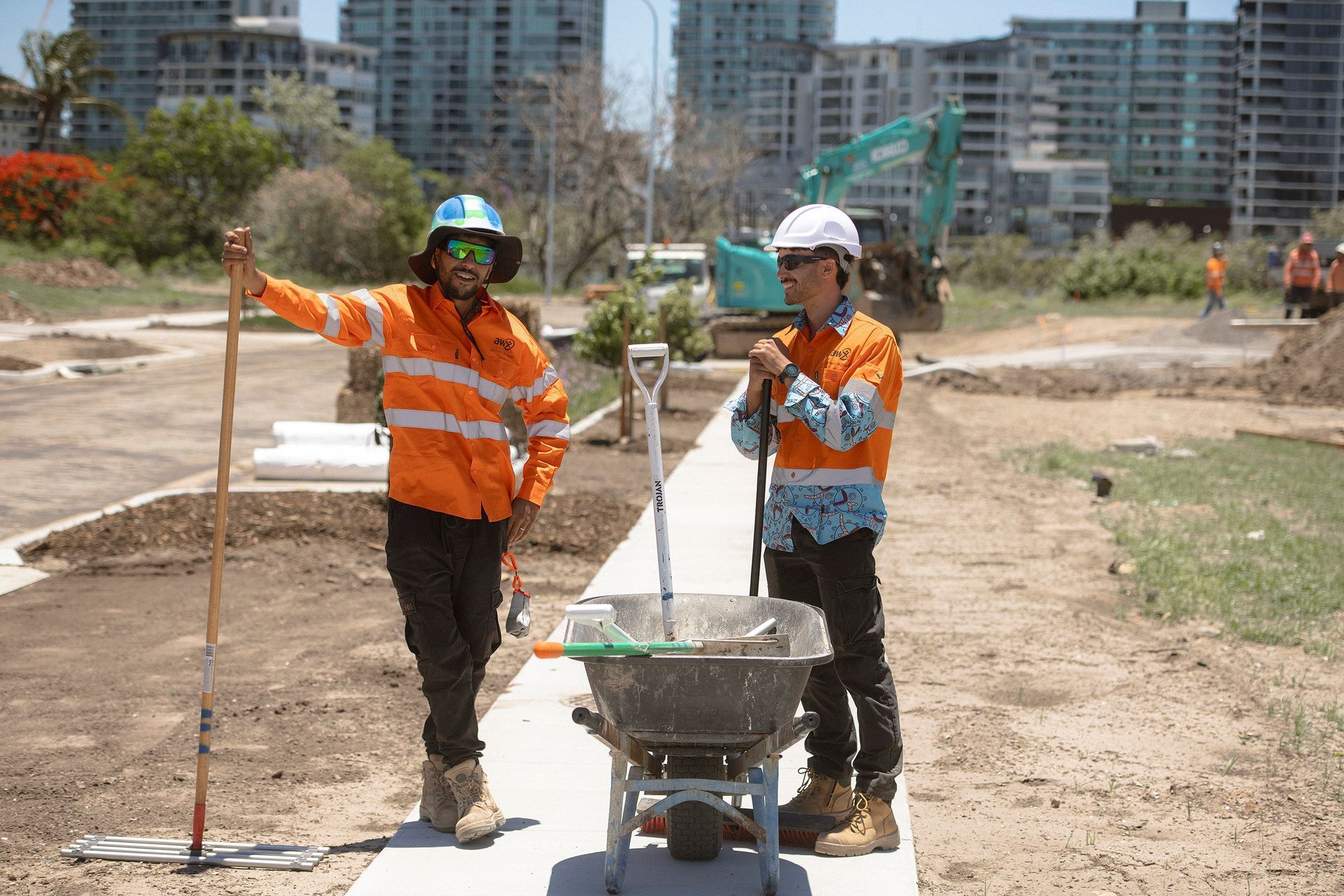 Two construction workers in orange safety gear stand beside a wheelbarrow on a dirt road at a building site.