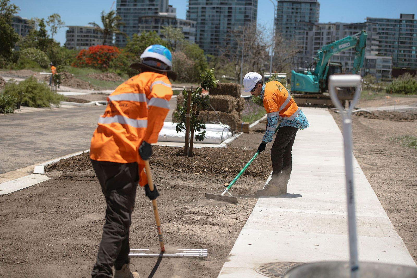 Two construction workers in orange vests raking dirt near a concrete pathway, with an excavator and buildings in the background.