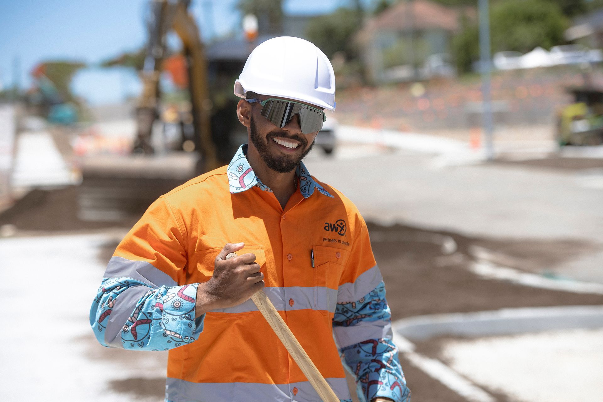 Construction worker in orange safety vest and hard hat smiles, holding tool, construction site in background.