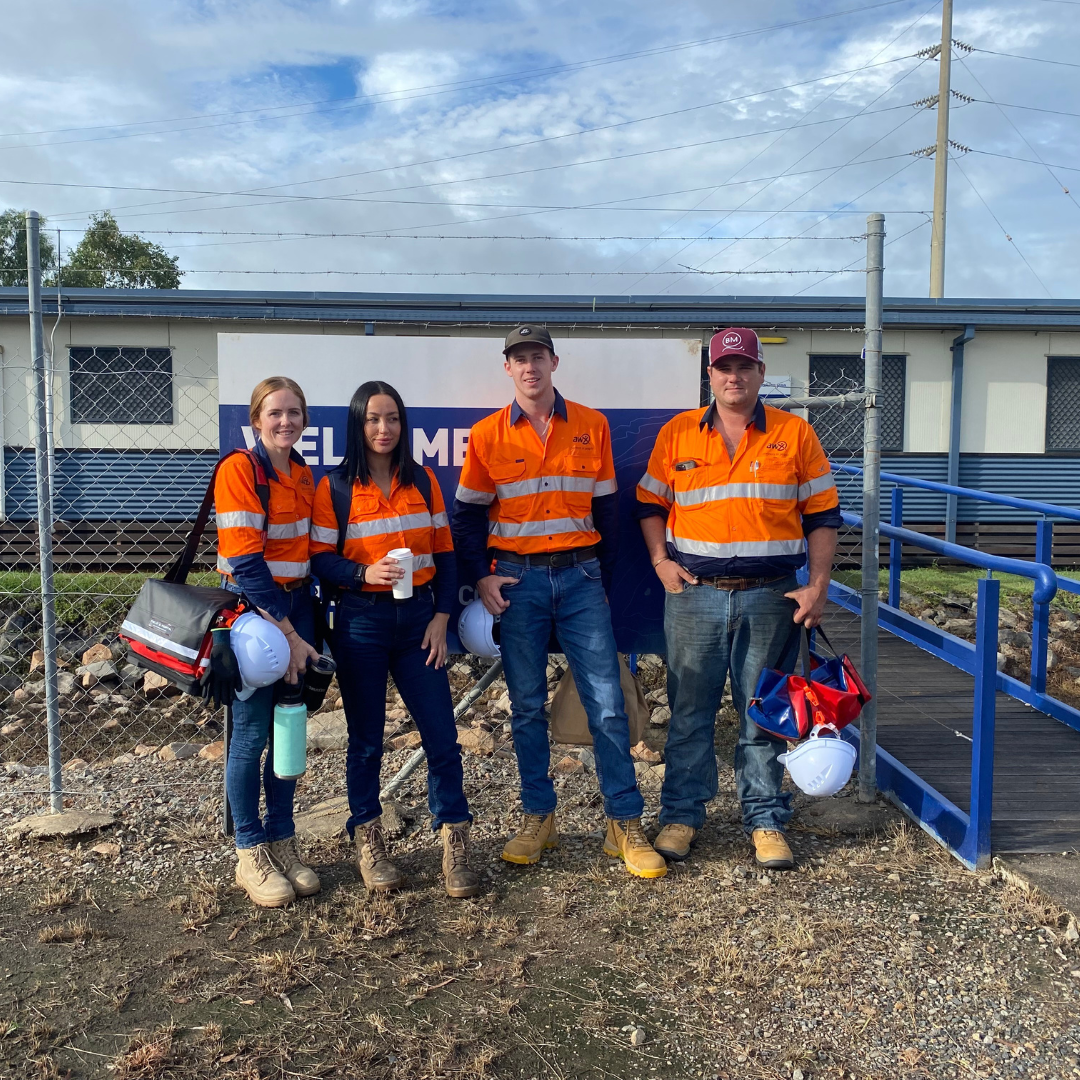 Four workers in orange safety shirts stand by a chain-link fence outside a worksite trailer