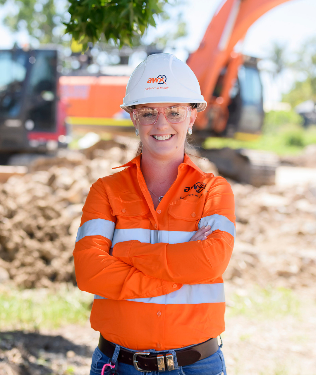 Woman in construction gear with arms crossed, smiling. Orange work shirt, white helmet, excavator in the background.
