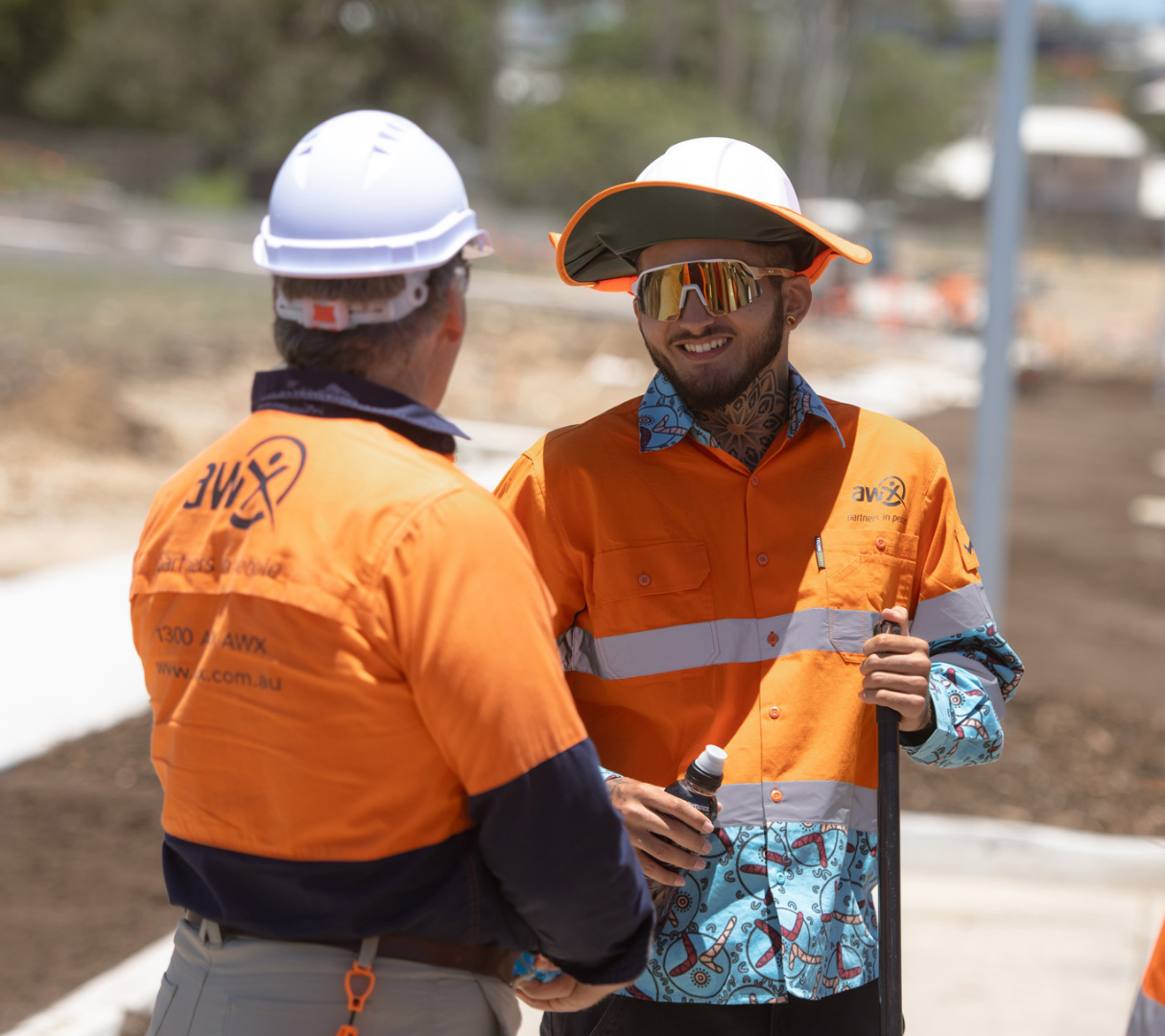 Two construction workers, one in white hardhat, the other in a hat and sunglasses, shake hands outdoors, both wearing orange safety shirts.