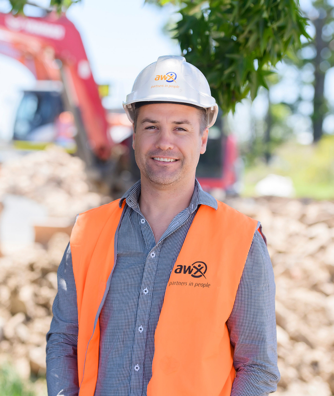 Man in construction workwear, arms crossed, smiling. Hard hat, excavator in background.