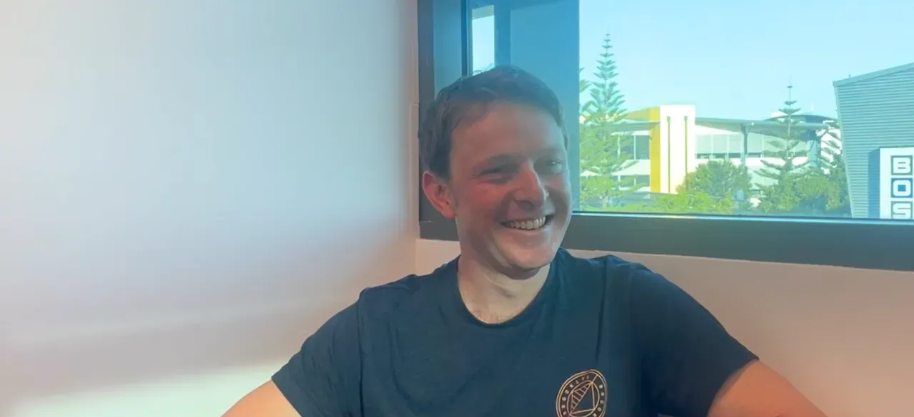 Smiling person in a dark shirt sitting indoors by a window with a scenic view outside