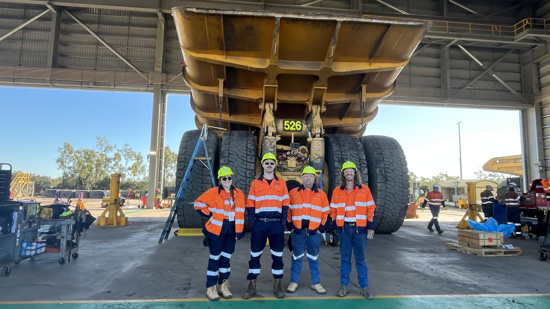 Five workers in safety gear stand under a massive dump truck in a maintenance bay.