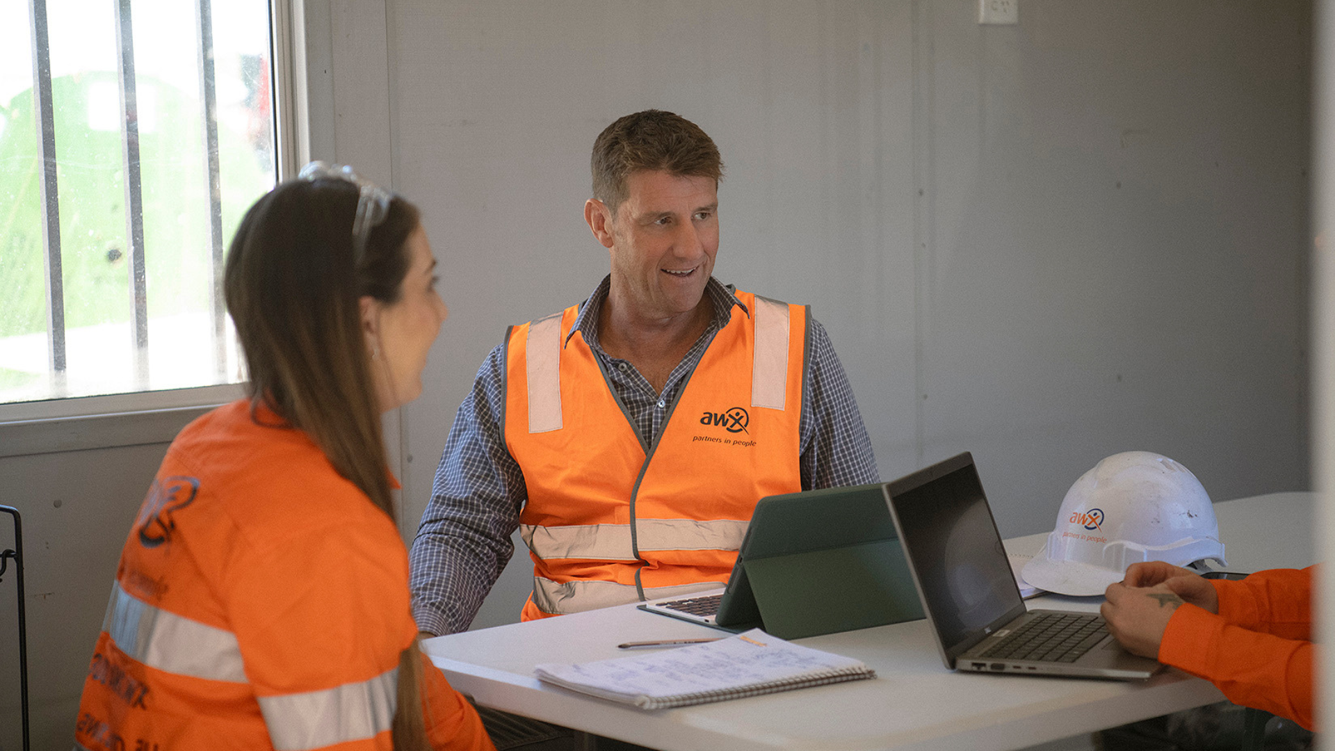 Construction workers in an office, one wearing a safety vest. They are sitting at a table with papers, a laptop and a tablet.