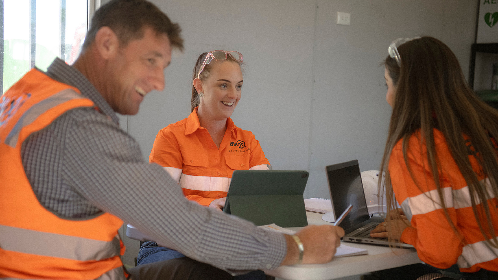 Three workers in high-visibility orange vests meeting around a laptop at a table indoors