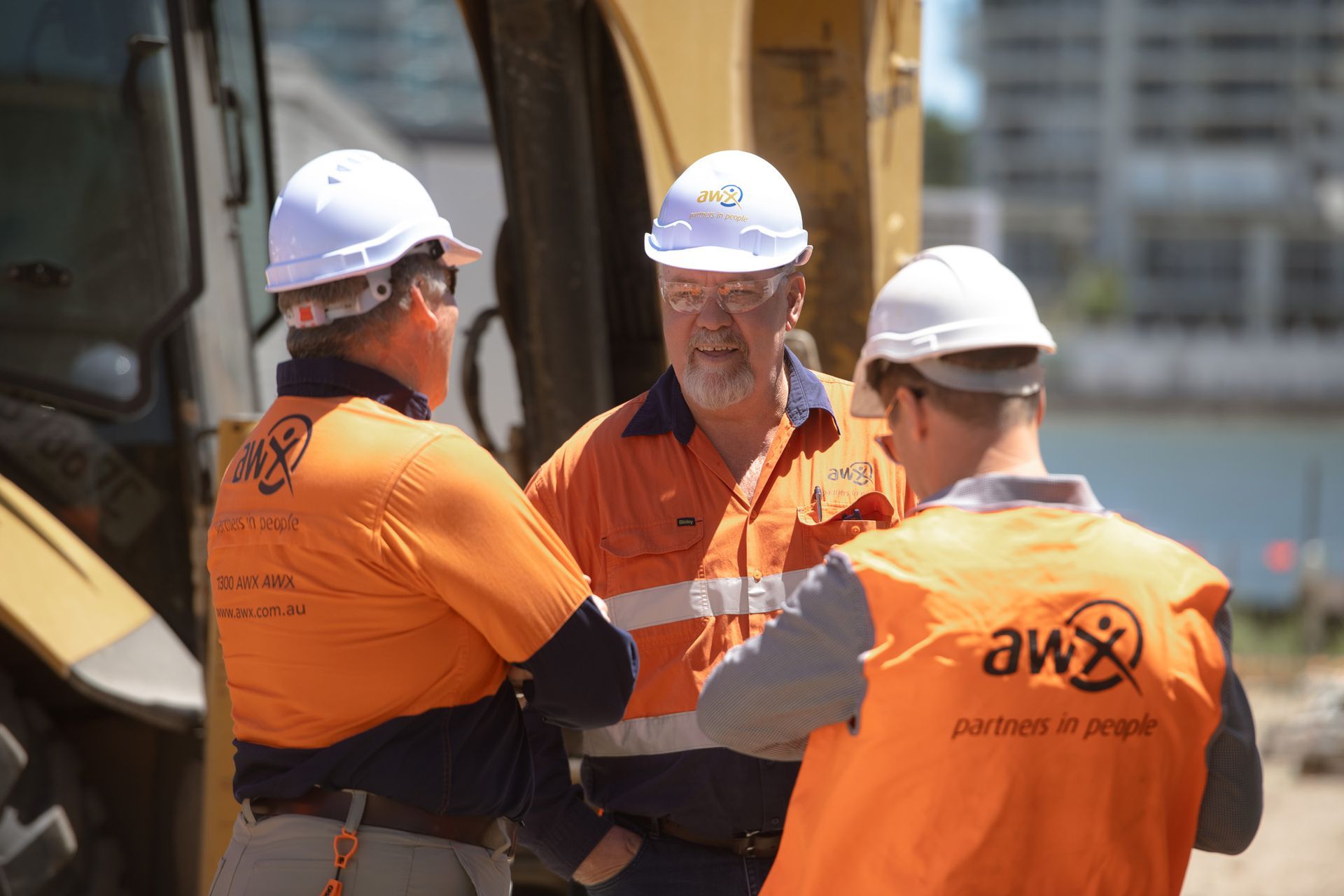 Three construction workers in hard hats and orange vests, talking near machinery.