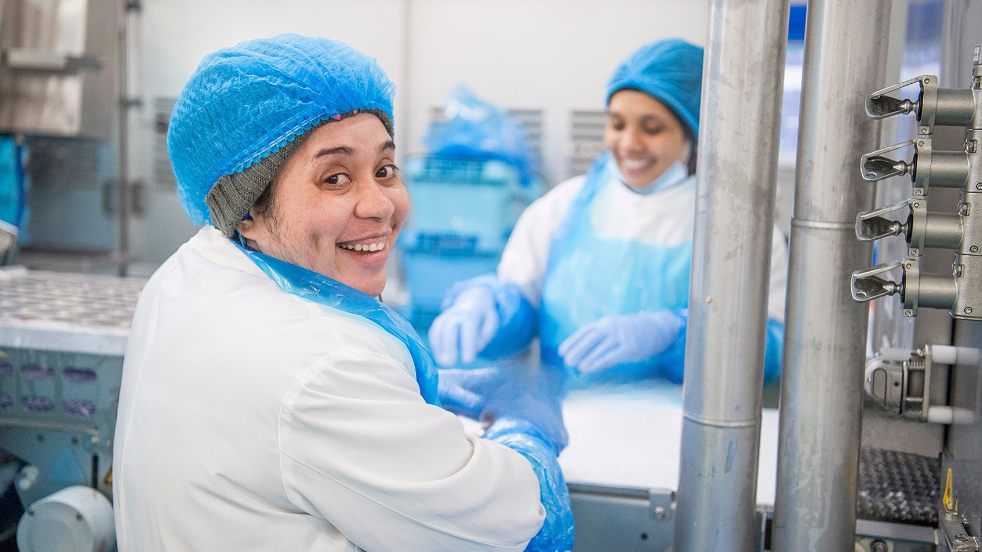 Two factory workers in blue hairnets smiling in a food processing facility