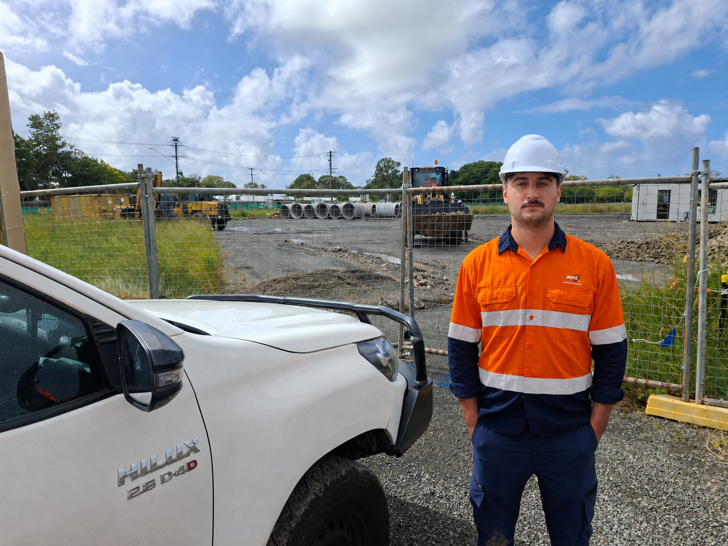 Worker in orange safety shirt and hard hat stands beside a white utility truck at a construction site.