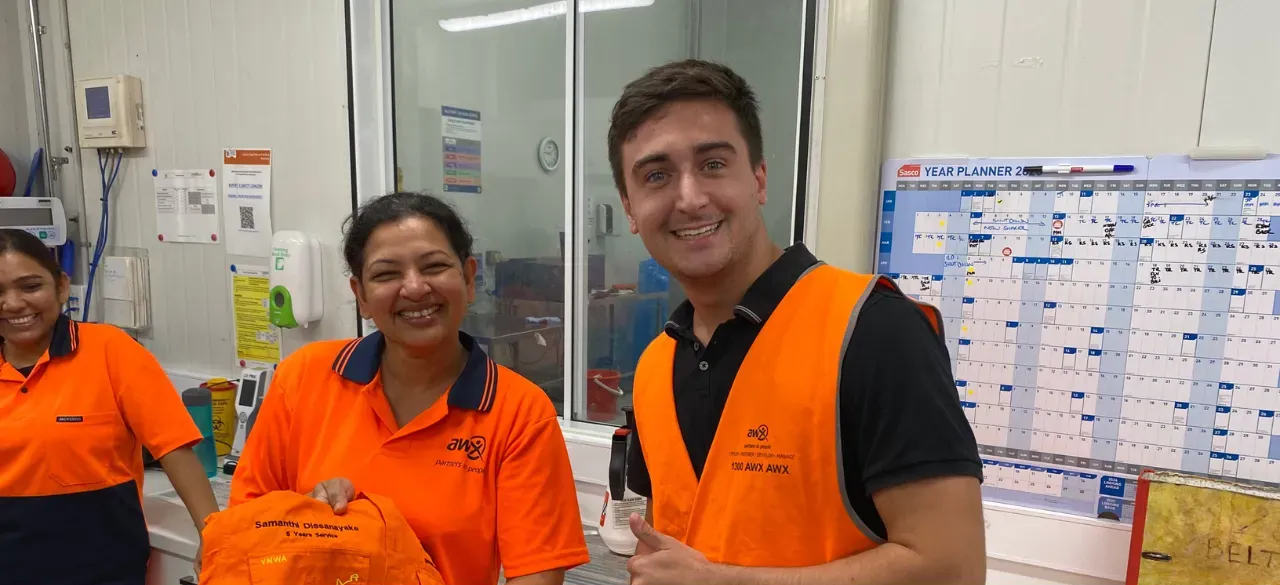 Three workers in orange safety vests standing in an office, smiling at the camera.