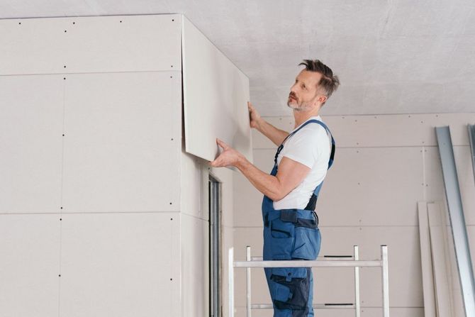 Person installing drywall in a bright room, standing on a ladder and aligning a wall panel.