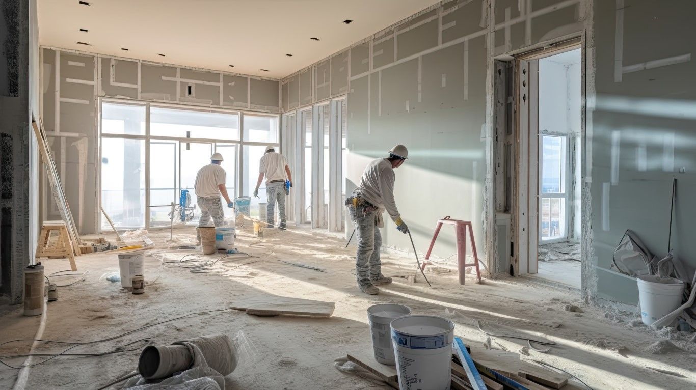 Workers installing drywall in a bright, unfinished room with tools and debris on the floor.