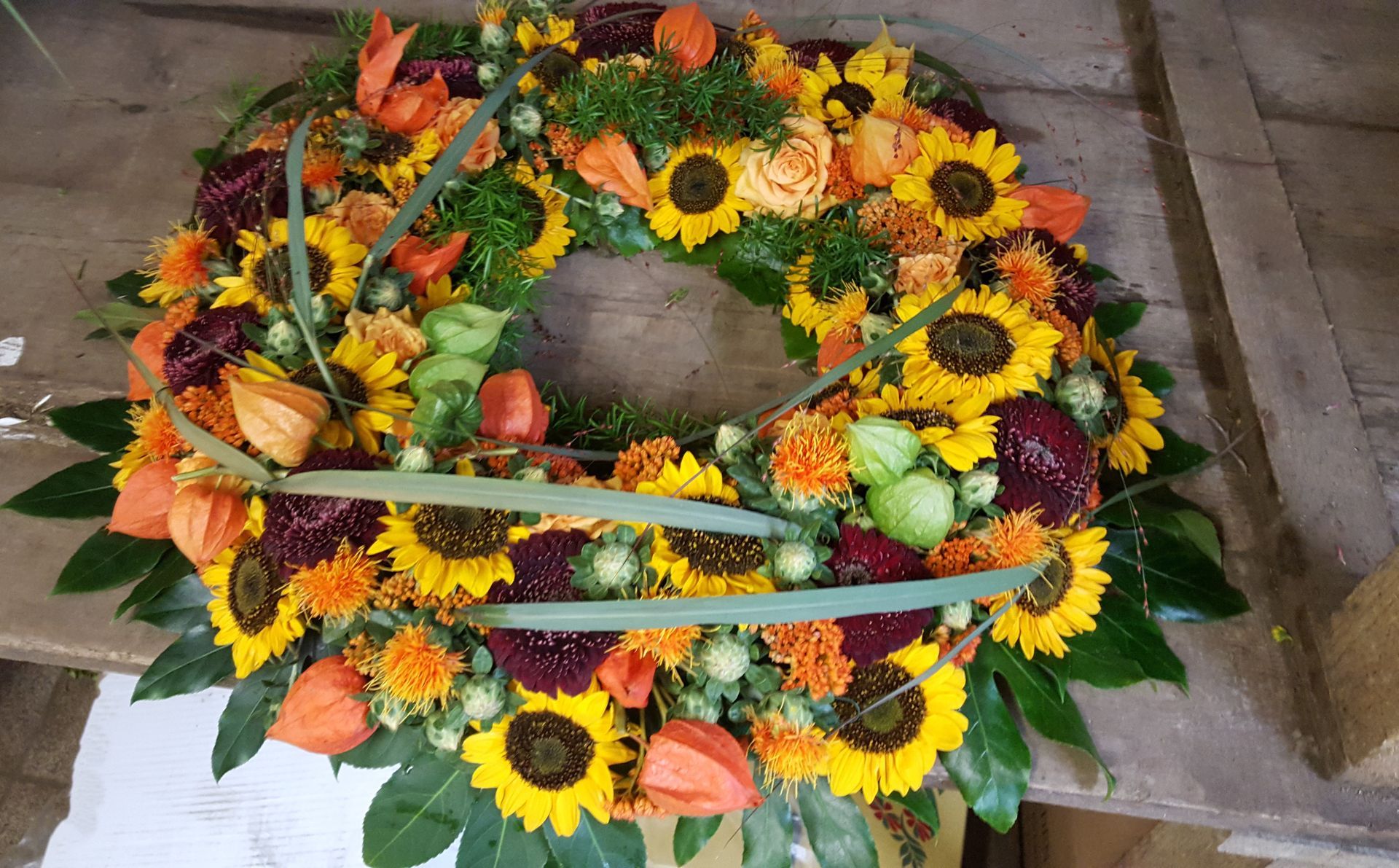 Auf einem Holztisch steht ein Kranz aus Sonnenblumen und anderen Blumen.