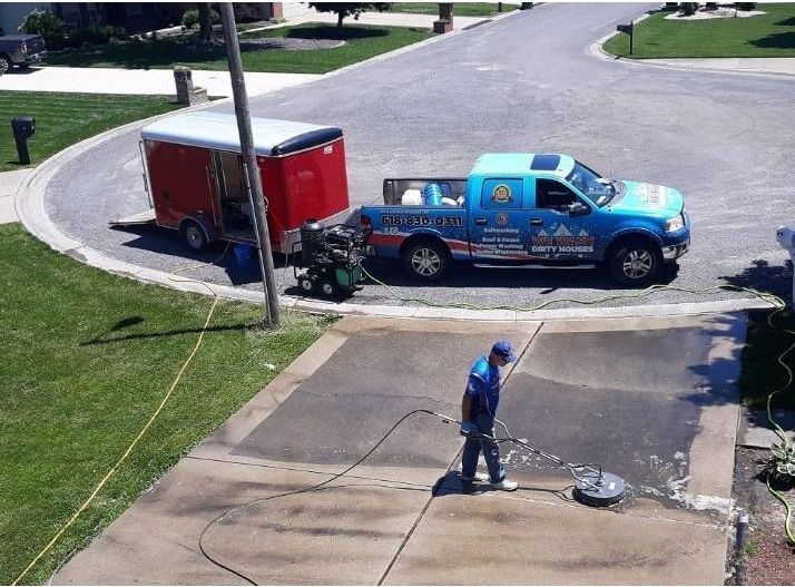 Worker pressure-washing driveway beside a blue pickup and red trailer on a suburban street.