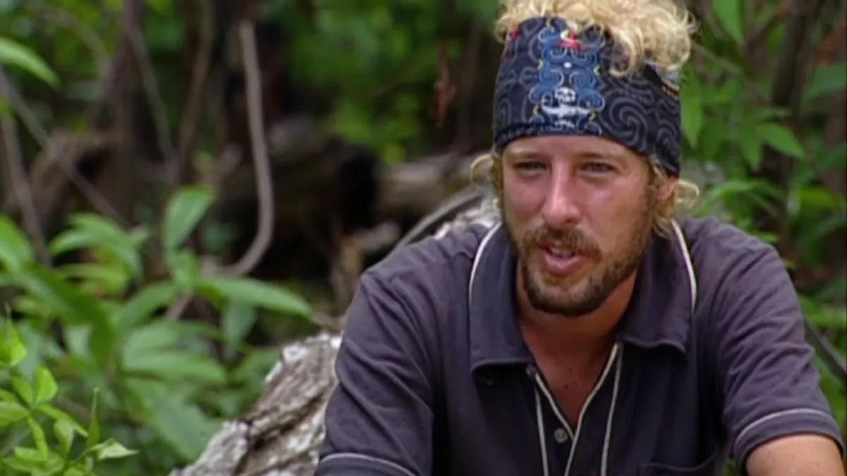 A man with a beard and a headband is sitting in the woods.
