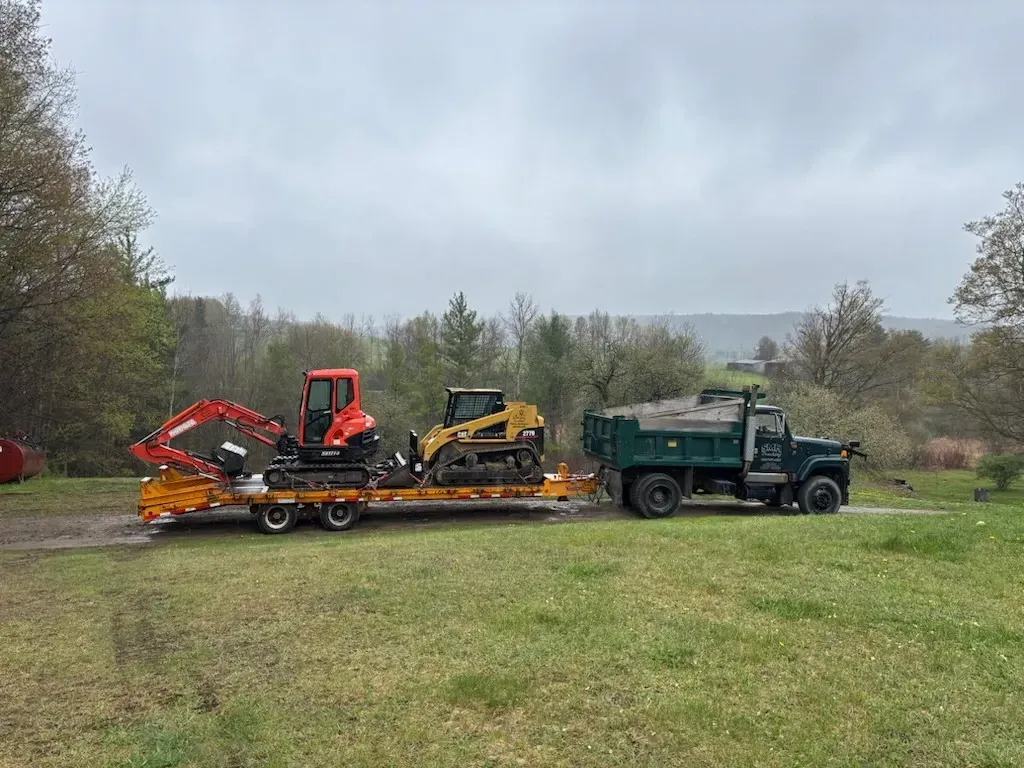 Green flatbed truck hauling two mini excavators on a trailer in a grassy field under a cloudy sky