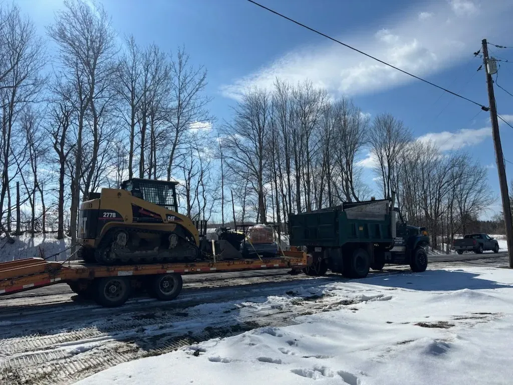 Two construction vehicles on a flatbed trailer in a snowy yard under bare trees