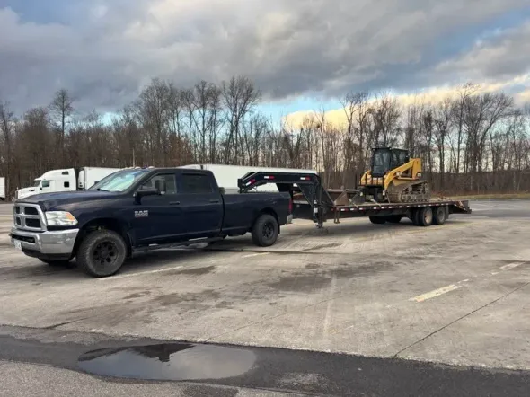 Black pickup truck towing a flatbed trailer with a yellow excavator on a lot under cloudy skies