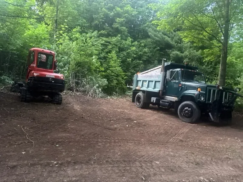Red excavator and green dump truck parked on a dirt clearing in a wooded area