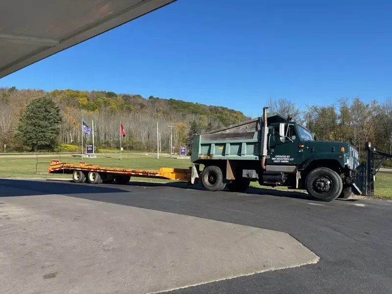 Green dump truck hauling a flatbed trailer in a parking lot under a blue sky