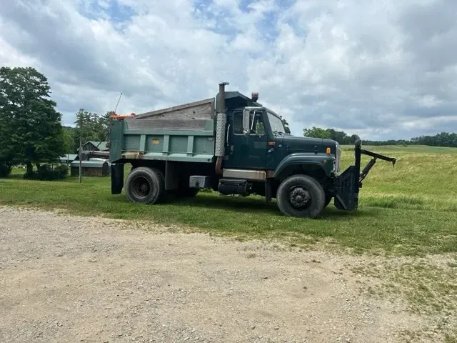Green dump truck parked on grass under cloudy sky, with a front-mounted plow.