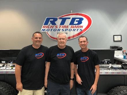 Three men are standing in front of a sign that says rtb rich 's tire barn motorsports