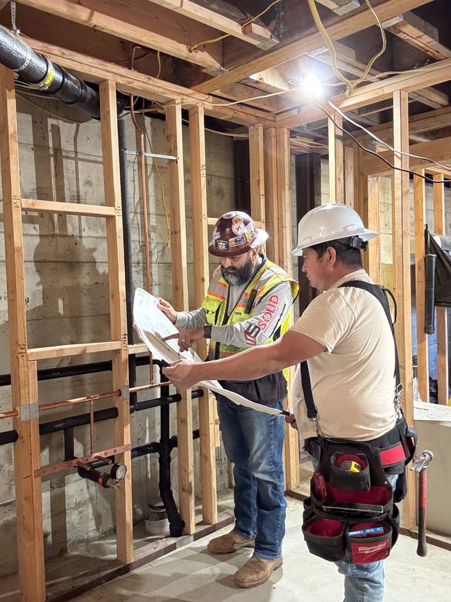 Construction workers building a wooden frame. They wear helmets, use tools, and work outdoors on a sunny day.