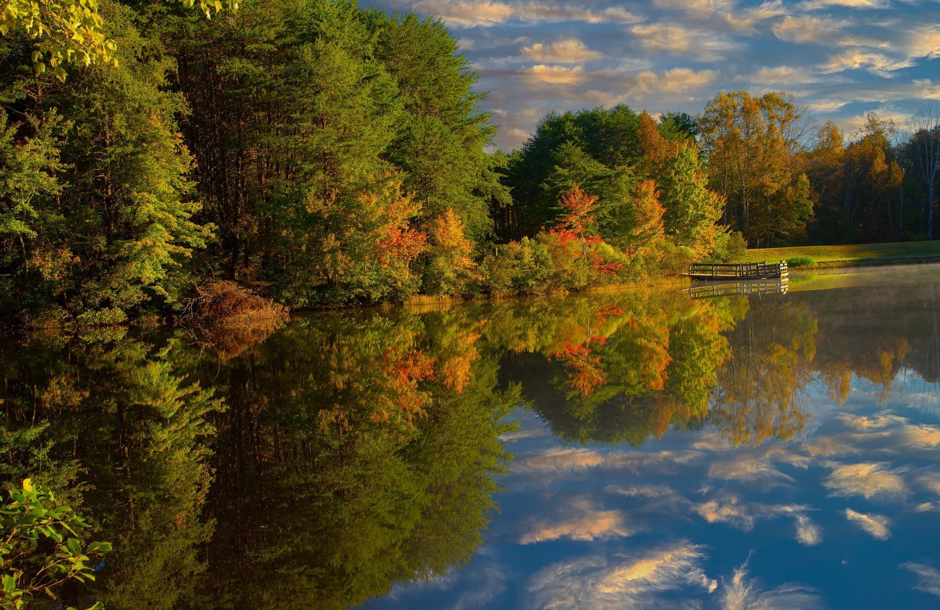 Lush green and autumn-colored trees reflect in calm water under a cloudy, blue sky.