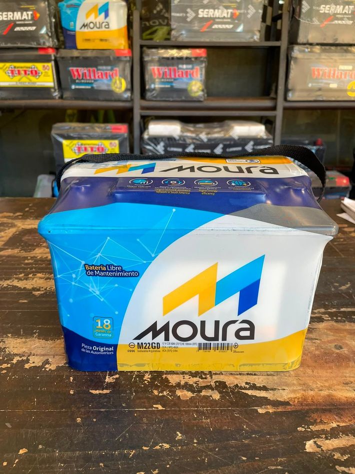 Una batería Moura azul y blanca sobre una superficie de madera, con más baterías apiladas detrás.