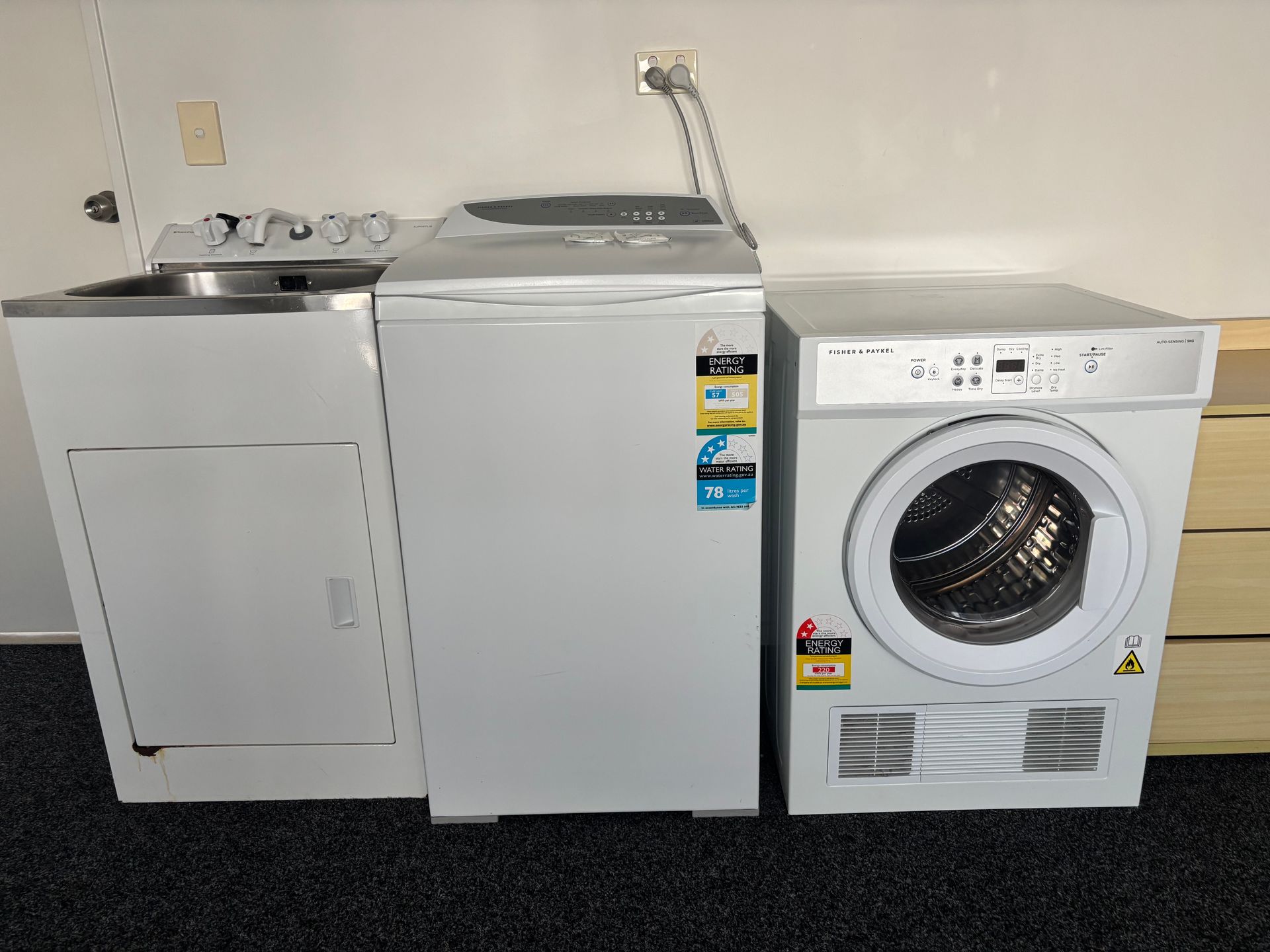 A laundry area with a washing machine, top-loading unit, and dryer aligned against a plain wall.