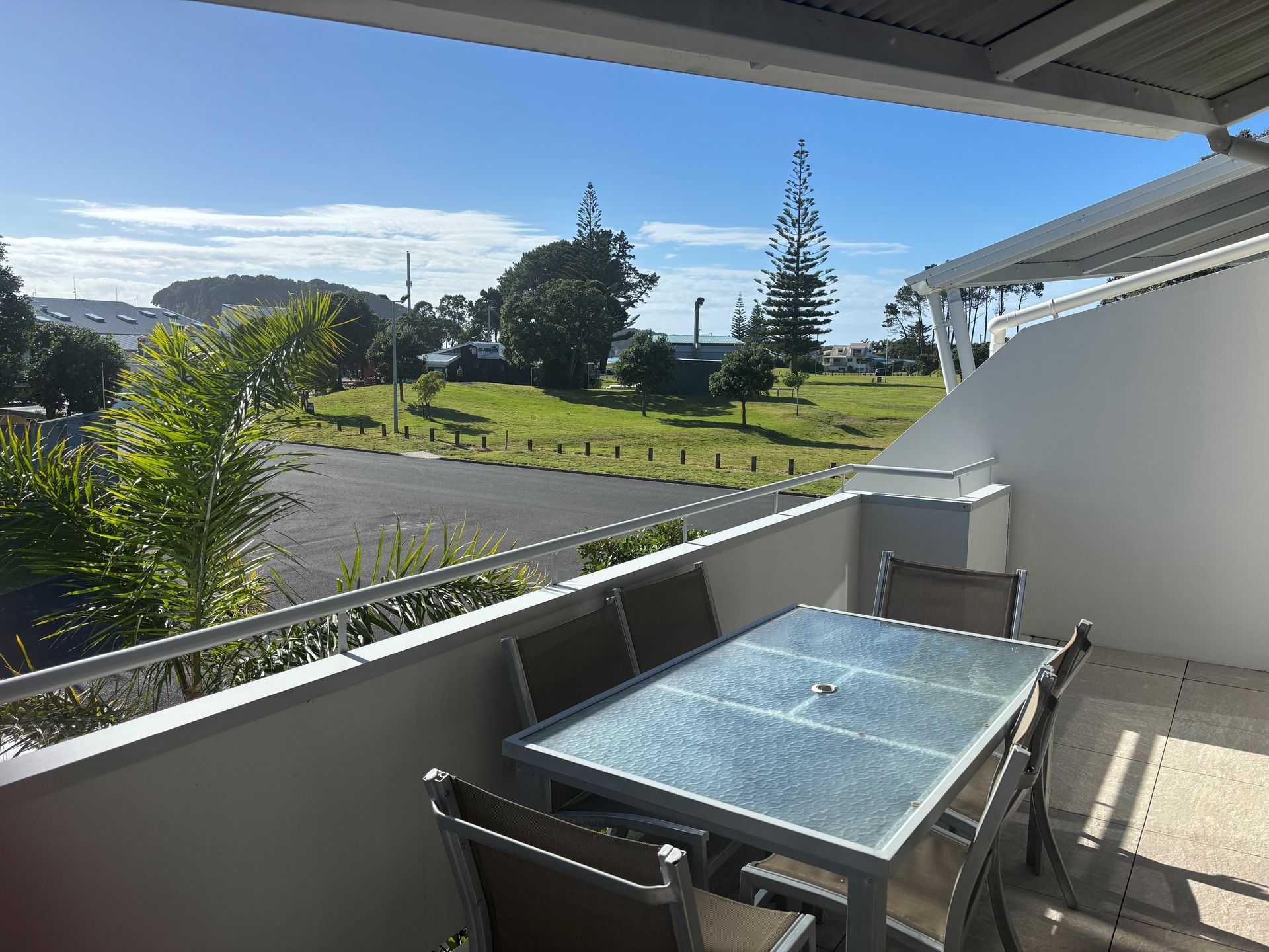 A glass-top outdoor dining table with chairs on a modern apartment balcony overlooking a grassy park and sunny blue sky.