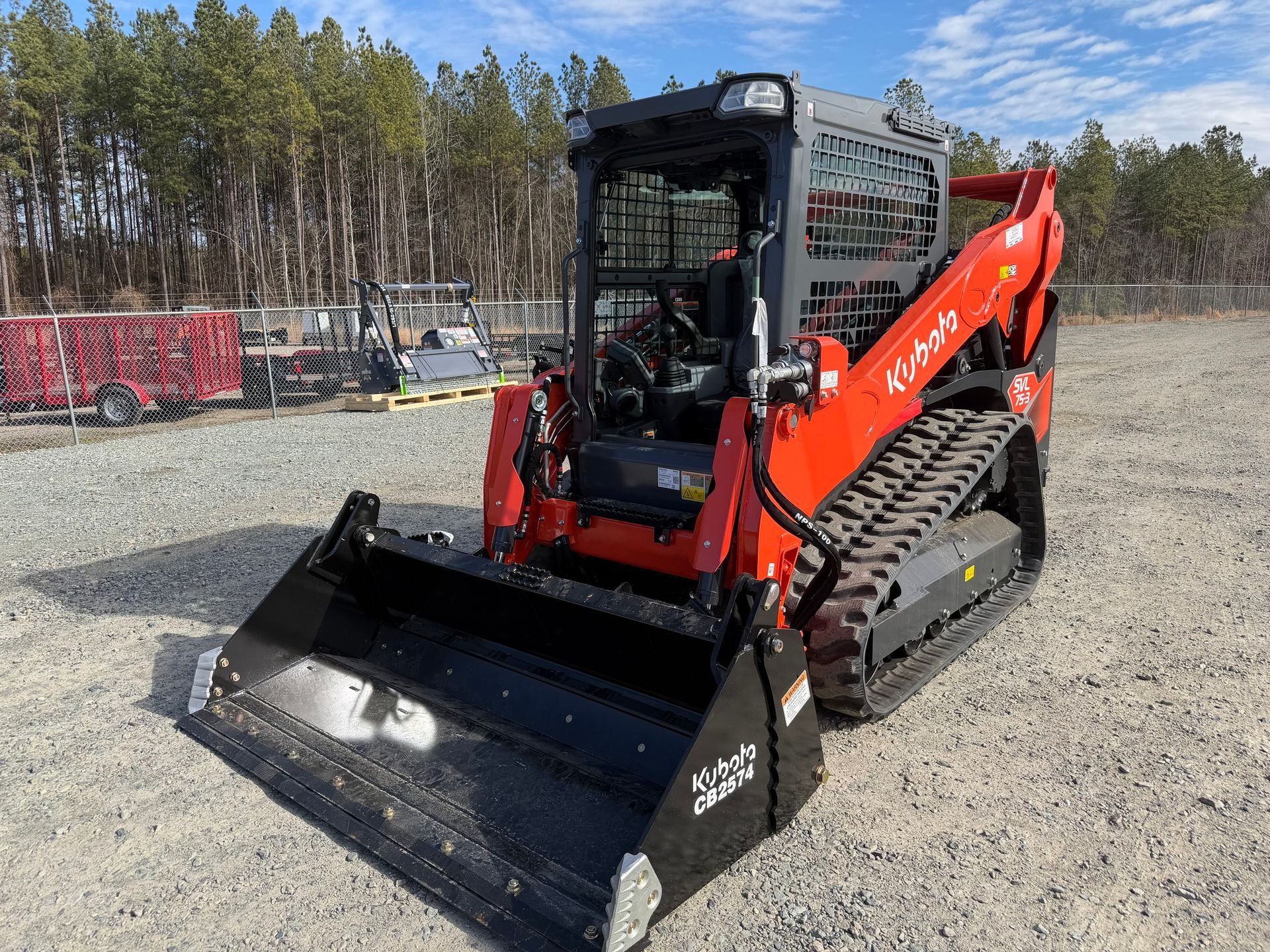 Red Kubota skid-steer loader with front bucket on gravel lot near trees