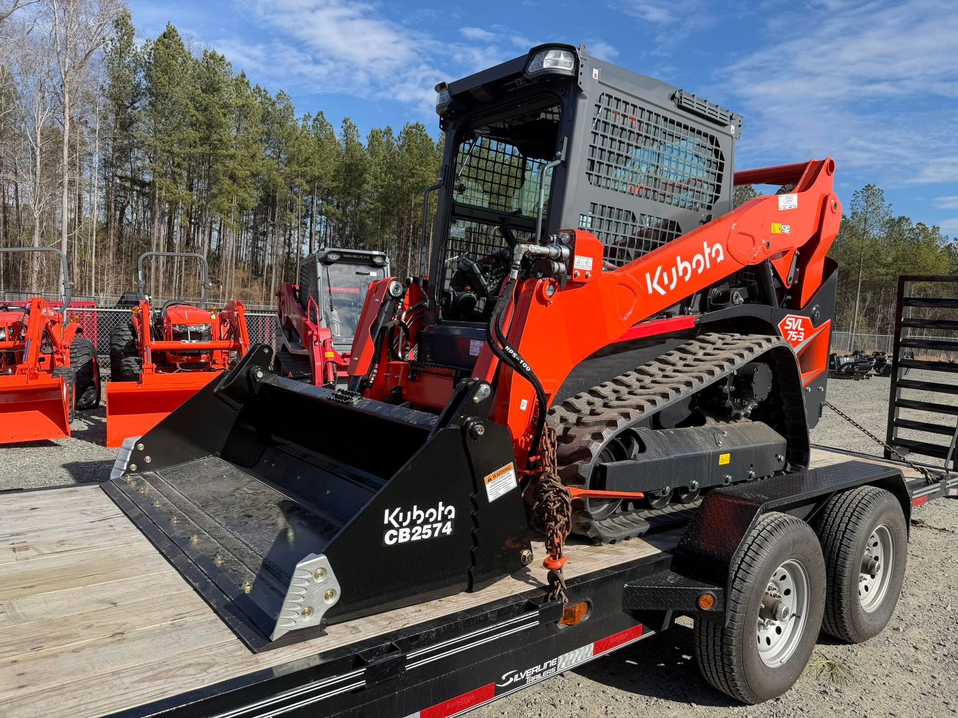 Orange Kubota skid steer loaded on a black trailer outdoors, with trees and blue sky in the background
