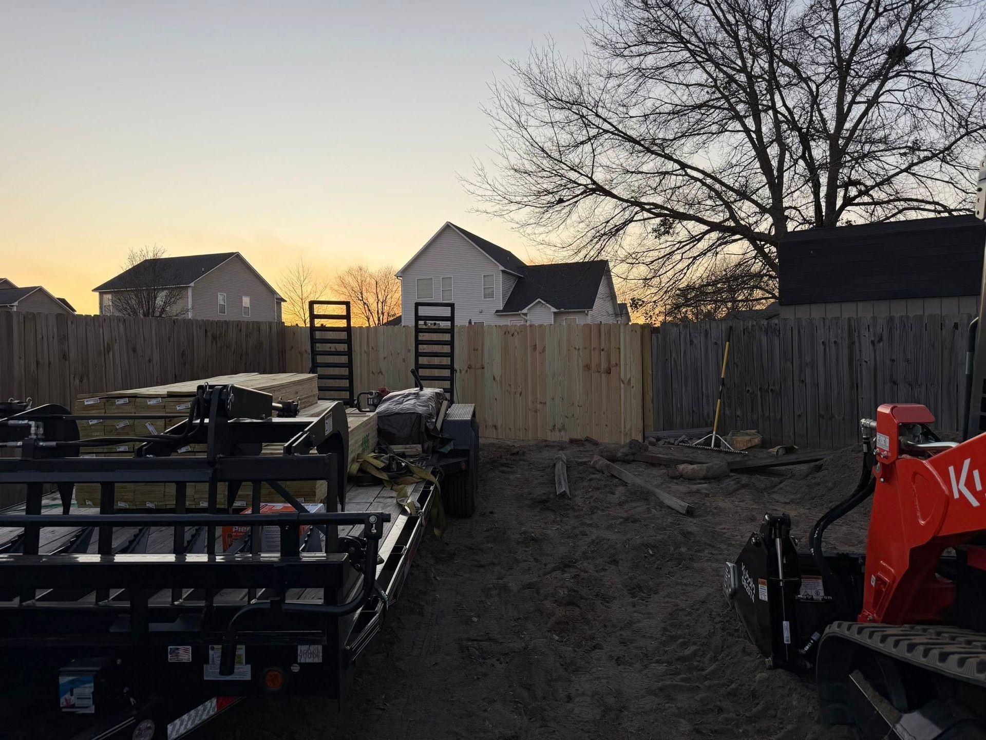 Construction equipment in a dirt lot at sunset, with houses and a fence in the background