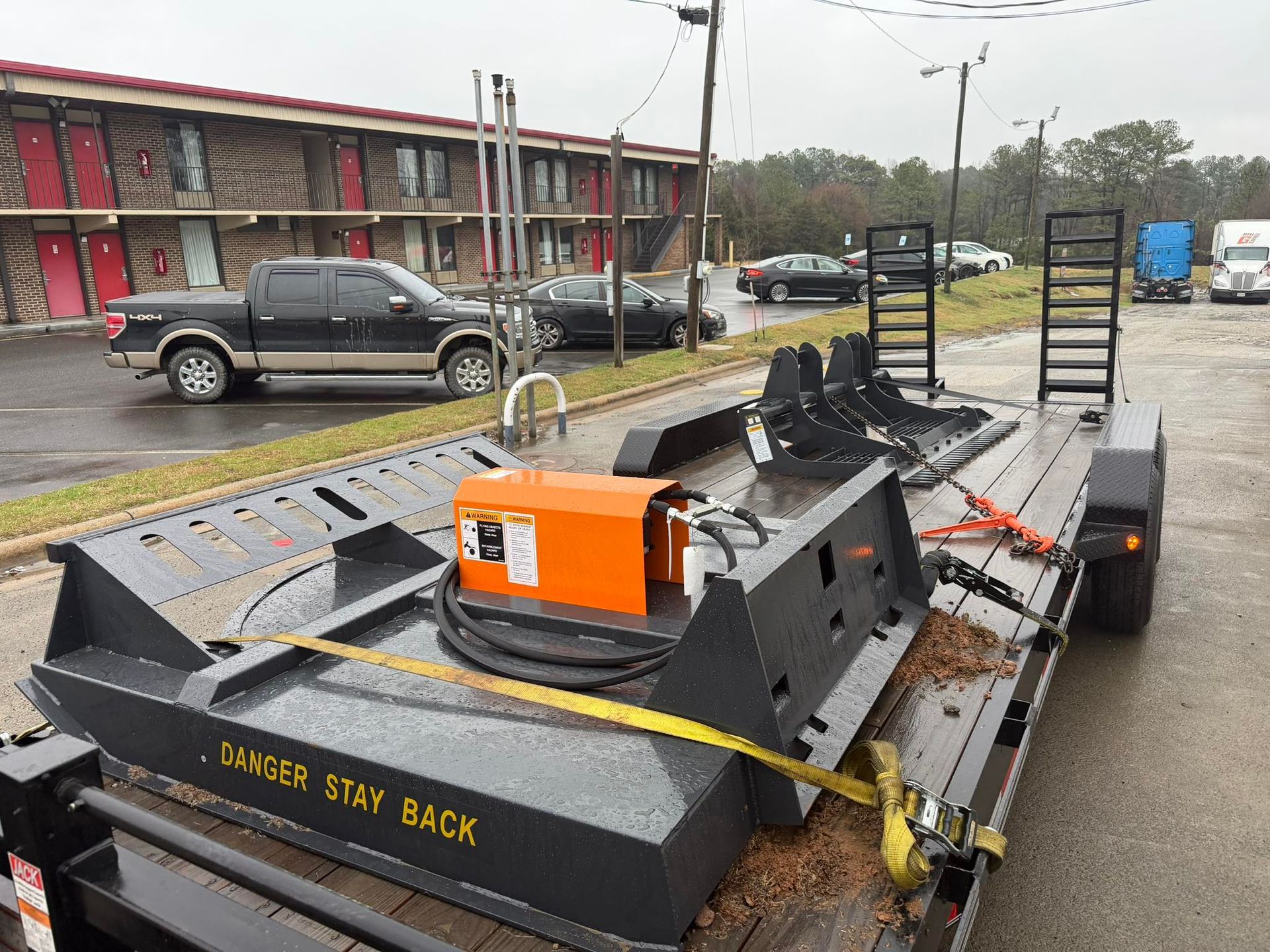Orange road-striping machine on a trailer in a parking lot with buildings and cars nearby