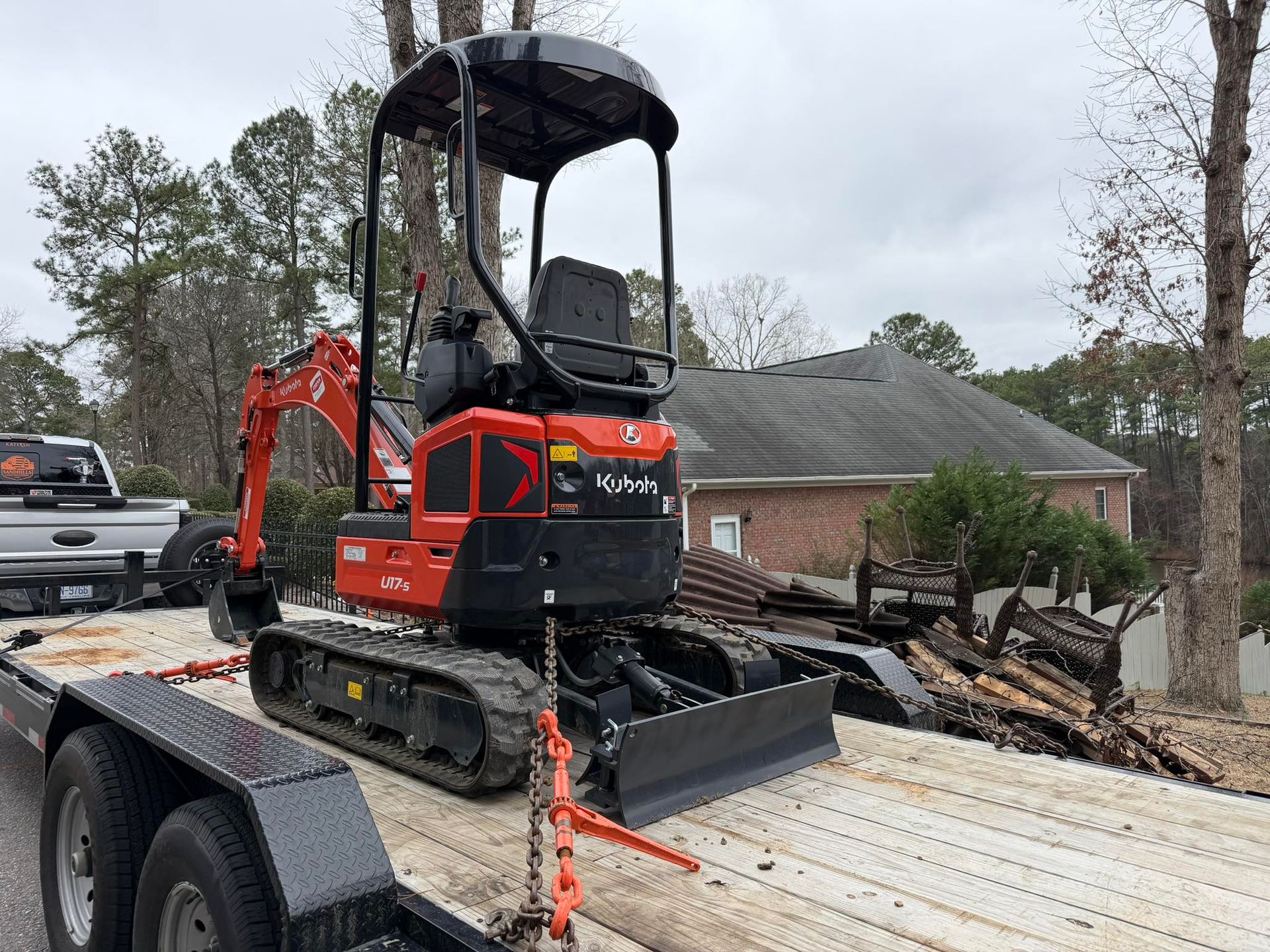 Red mini excavator strapped to a trailer amid storm debris in a damaged yard