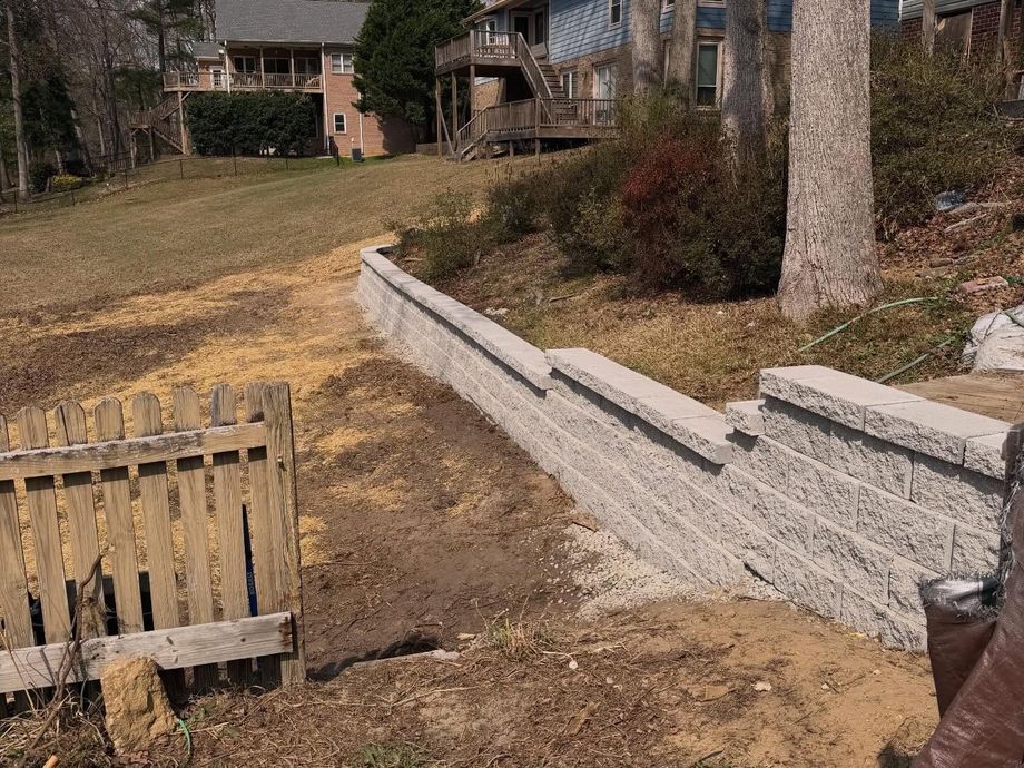 Backyard with a long concrete retaining wall, dirt slope, and houses in the background