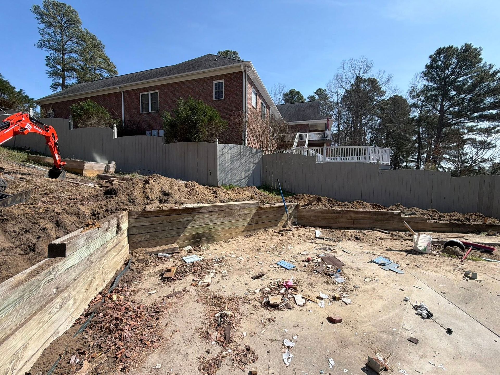 Construction site with excavator, dirt lot, and brick house behind a white fence