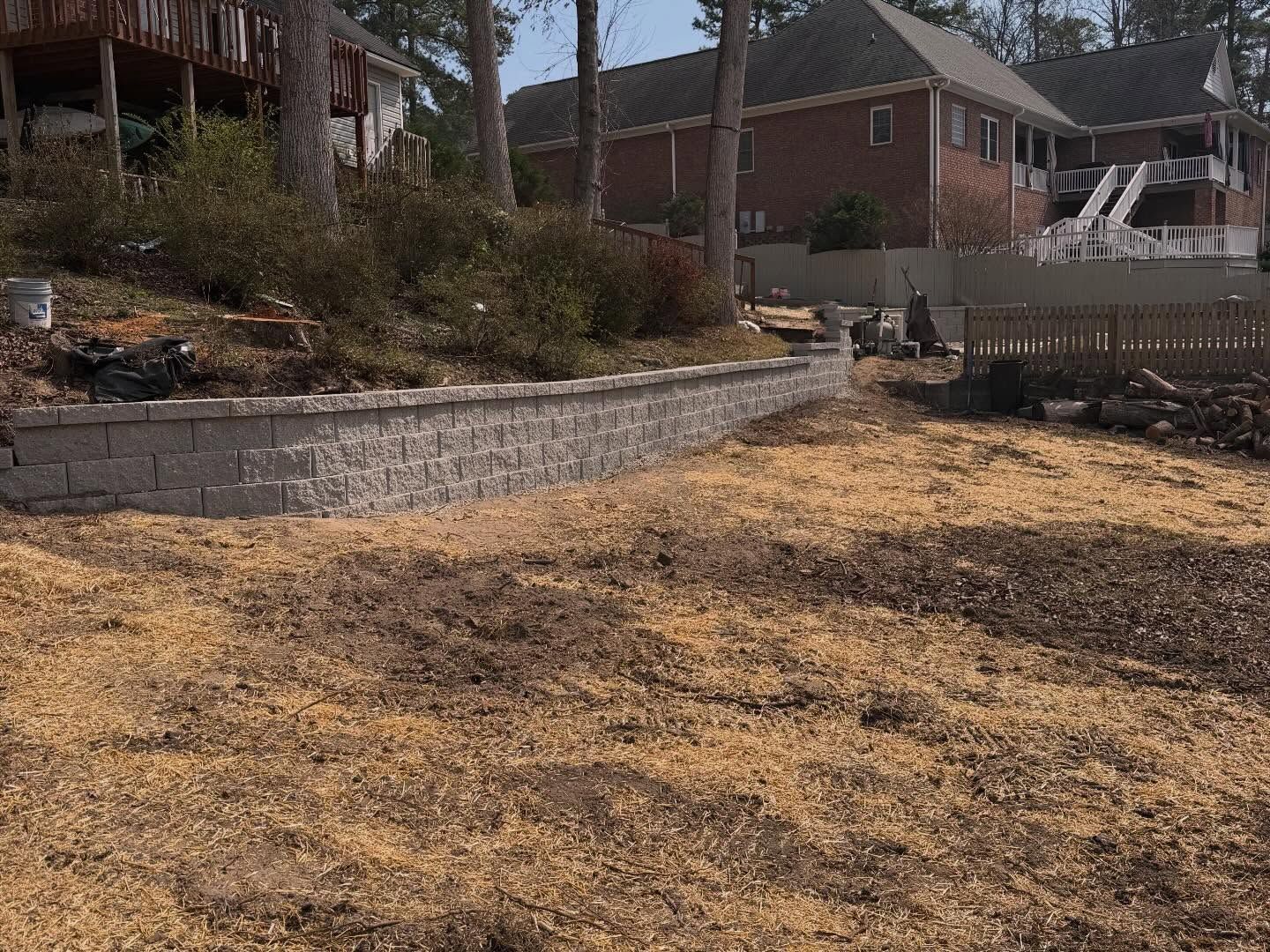 Dry backyard with straw-covered ground, stone retaining wall, shrubs, and brick houses in the background