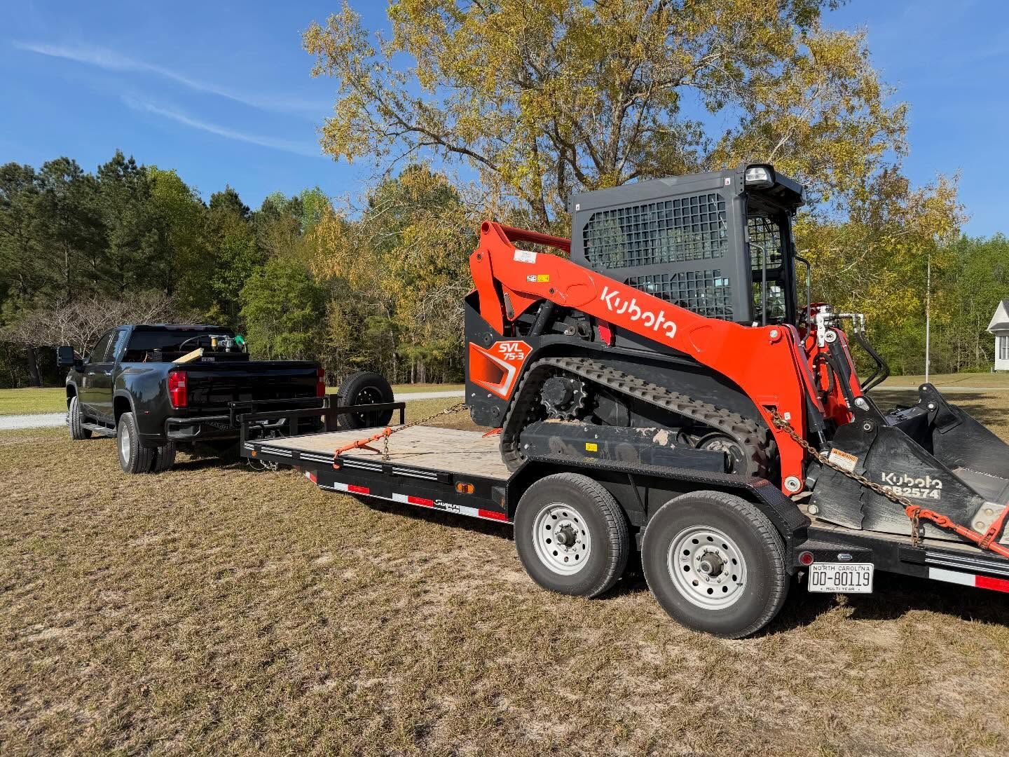 Orange Kubota skid steer on trailer being towed through a muddy field with a black truck, fall trees in background