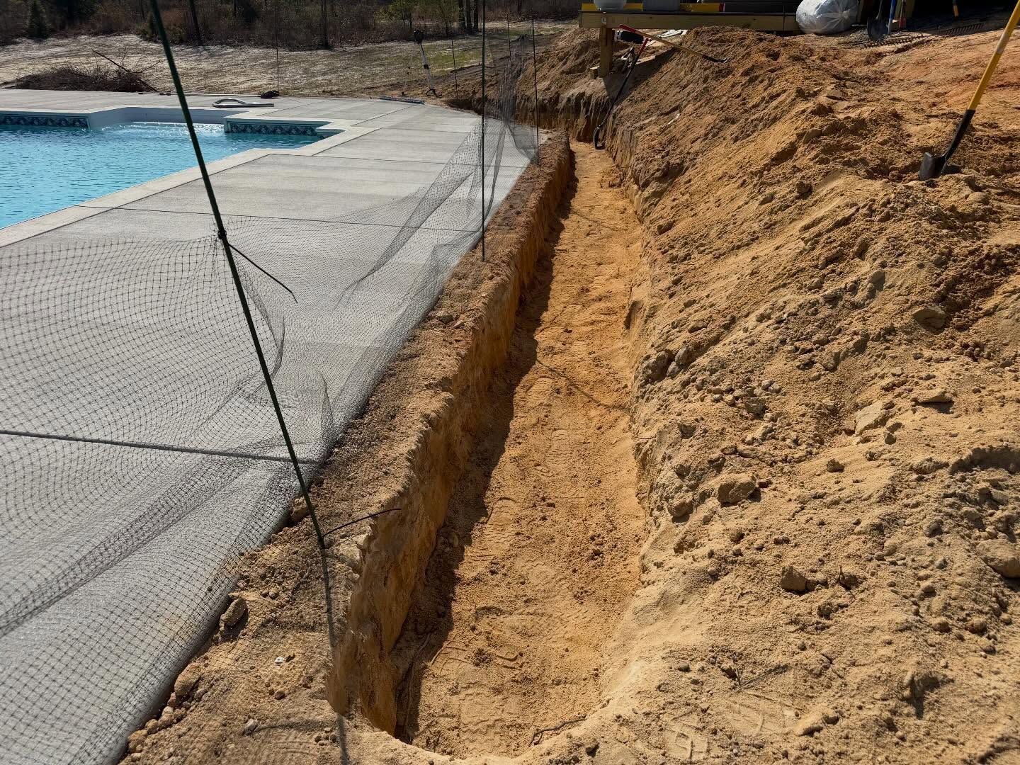 Excavated trench beside a swimming pool and concrete deck, with exposed soil and support stakes
