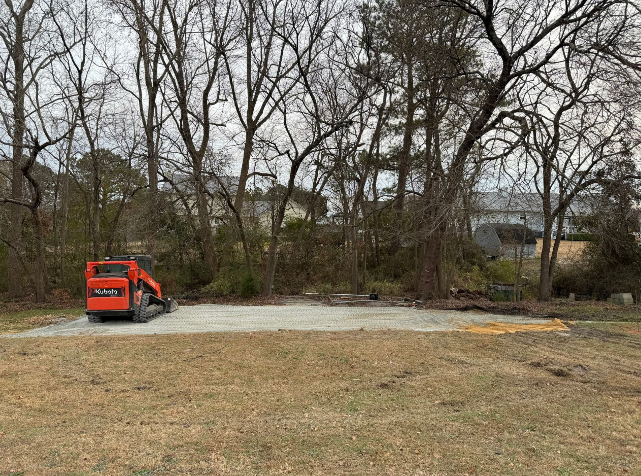 Small orange excavator on a gravel area beside leafless trees and brush