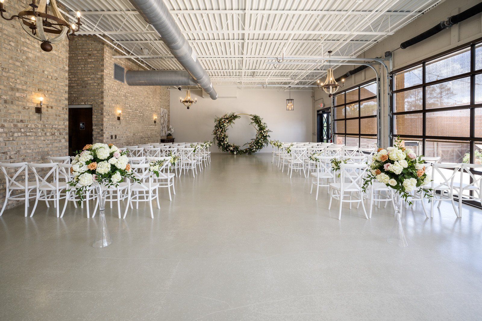 Wedding venue: white chairs, floral arrangements, large windows, and a circular floral arch.