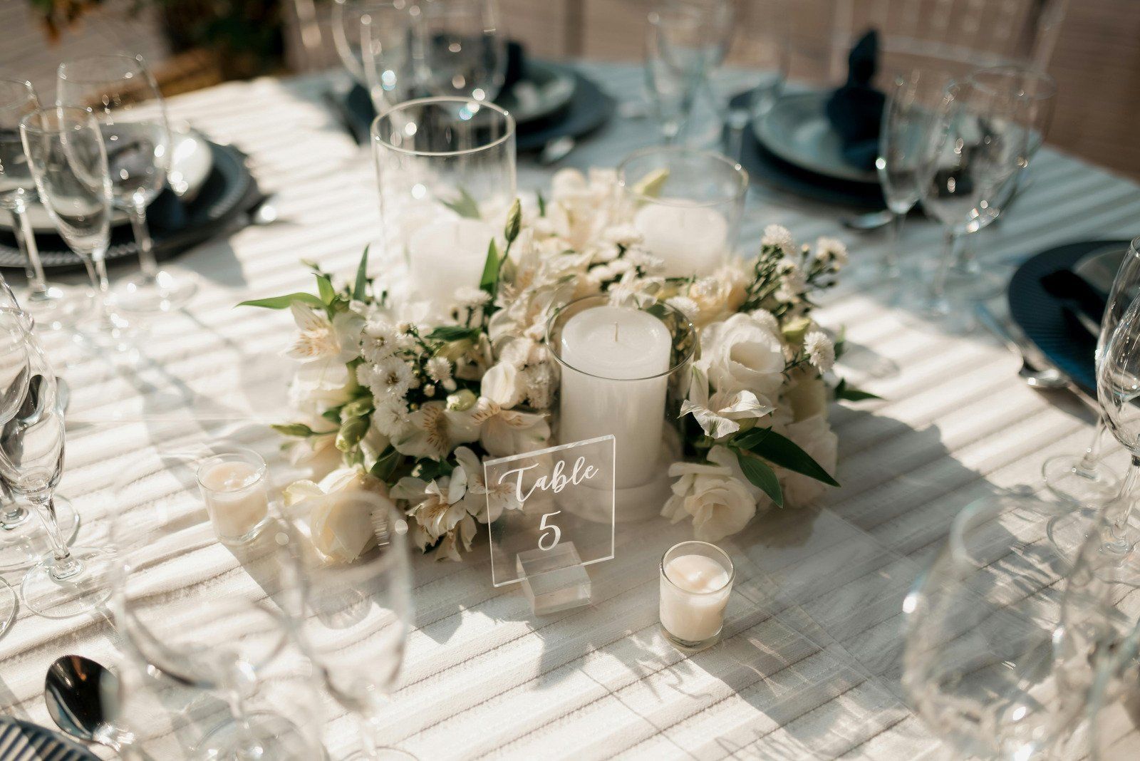 White floral centerpiece with candles on a table set for a formal event, with place settings and a table number.
