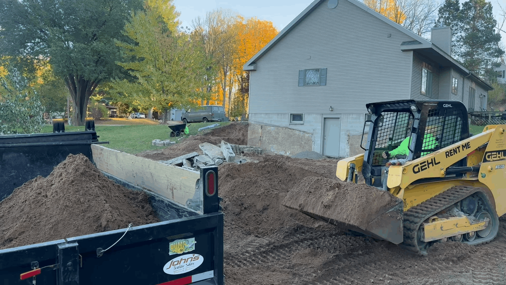 A yellow skid steer loads dirt into a dump truck near a house; outdoor construction.