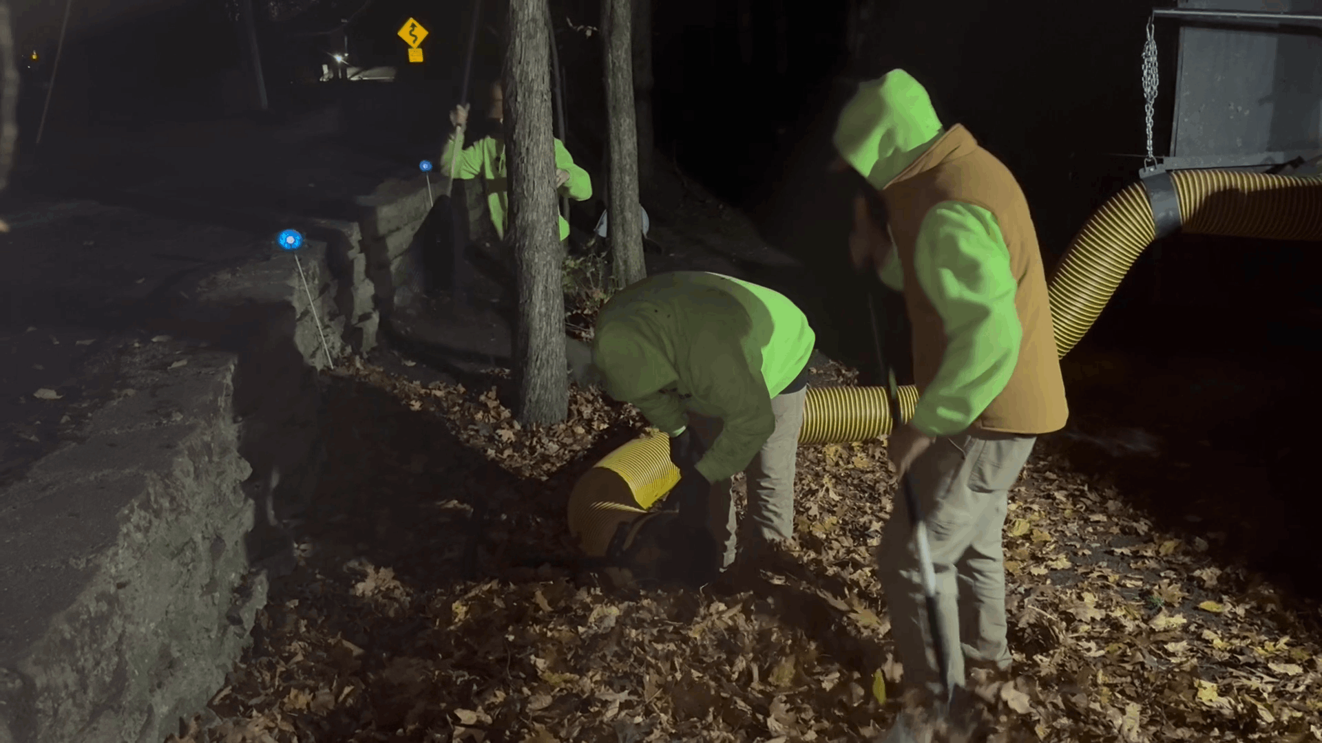 Three workers in safety vests and hoods using a vacuum hose to clean up leaves at night.