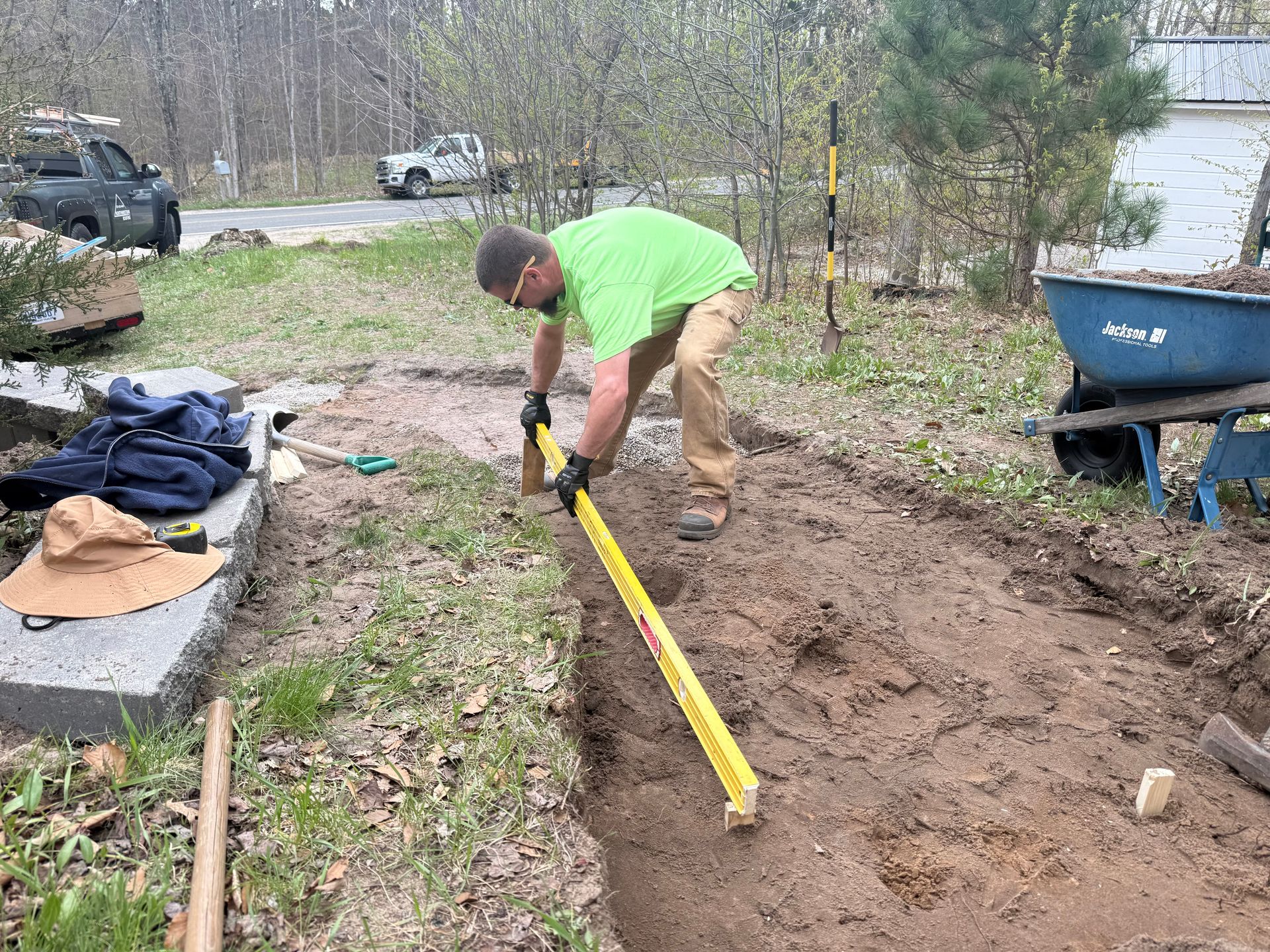 Person in a green shirt digging a garden trench with a shovel beside a wheelbarrow.
