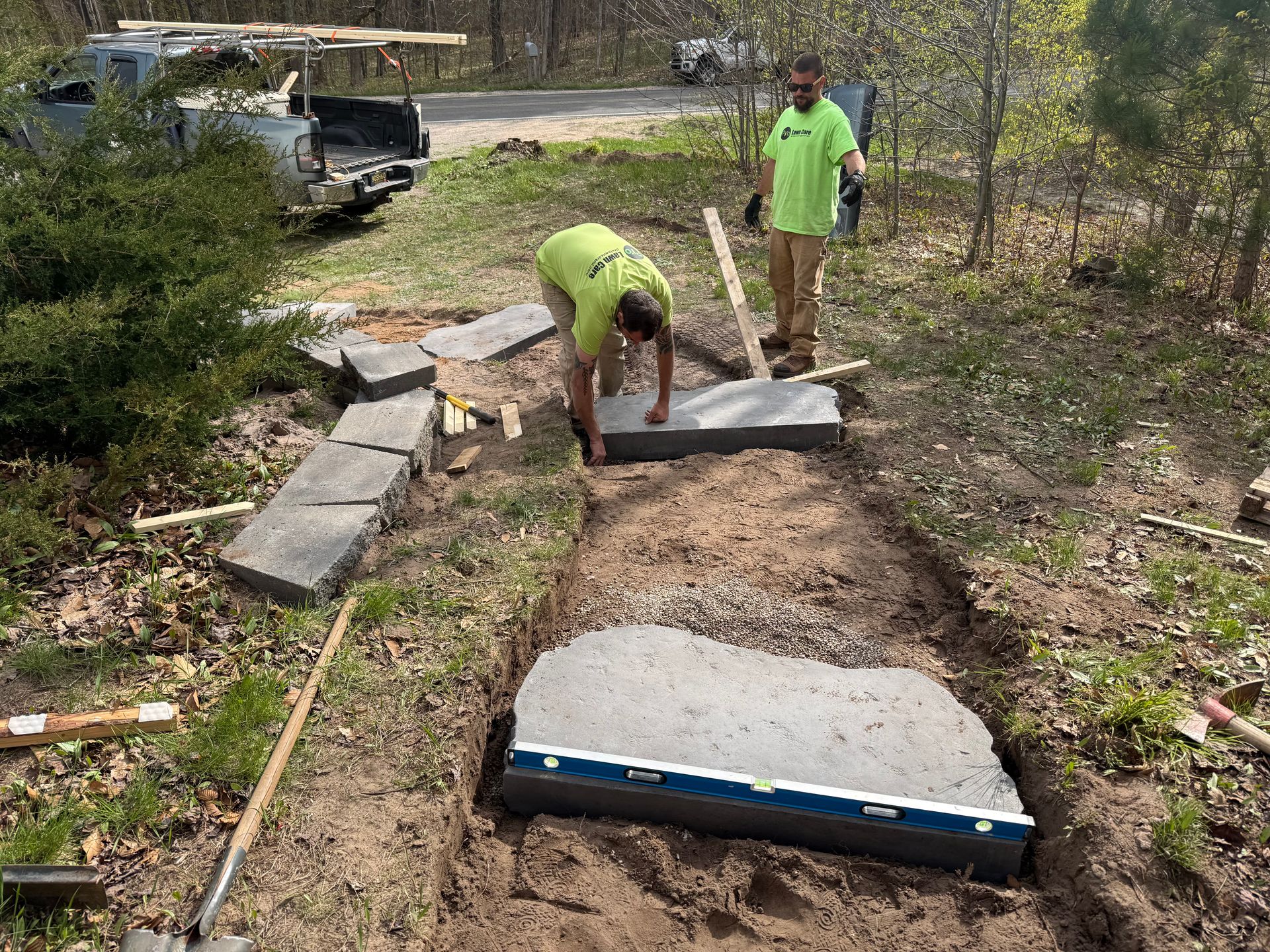 Two workers laying concrete pavers on a dirt path beside a yard and parked truck