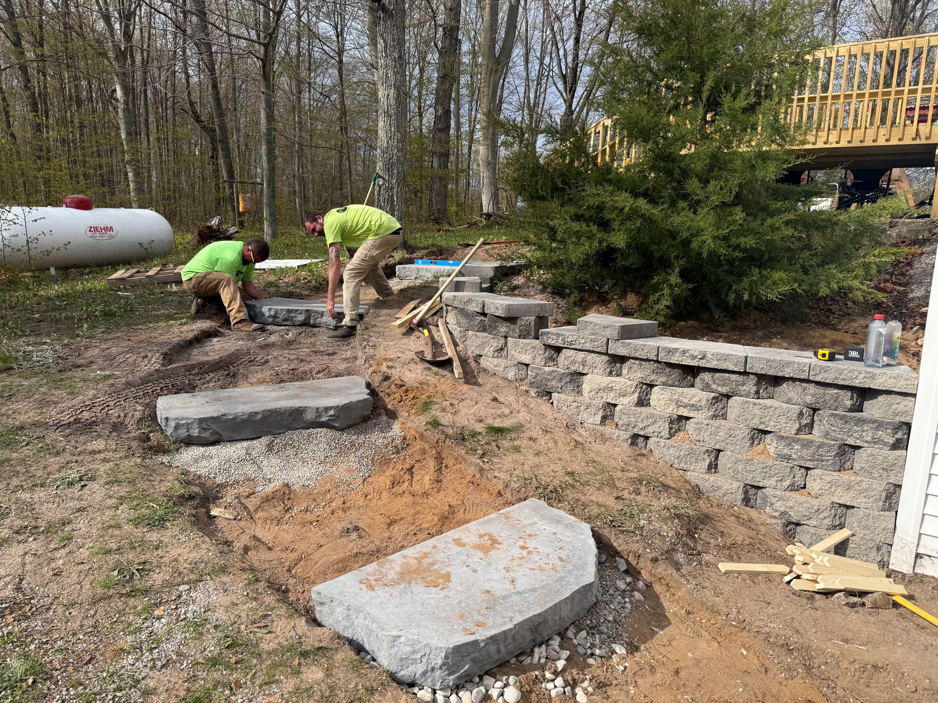 Workers building a stone retaining wall beside a backyard patio in a wooded yard